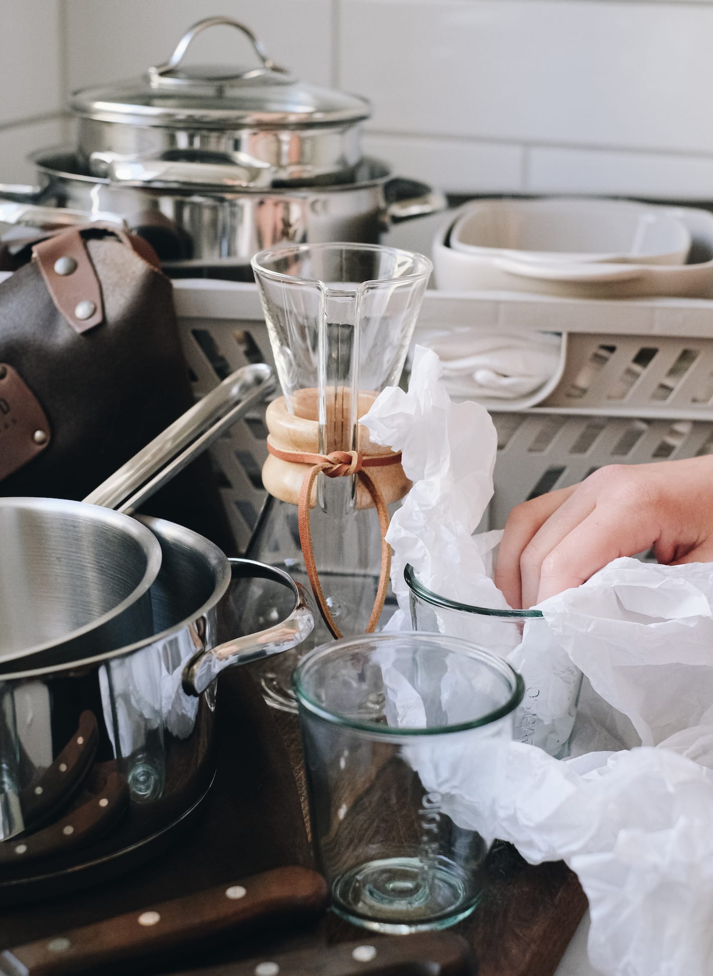 En person pakker inn glass fra Eva Solo i silkepapir. Ved siden av st&aring;r et grytesett og i bakgrunnen en vannkaraffel, og en plastkasse med ildfaste former og flere gryter.